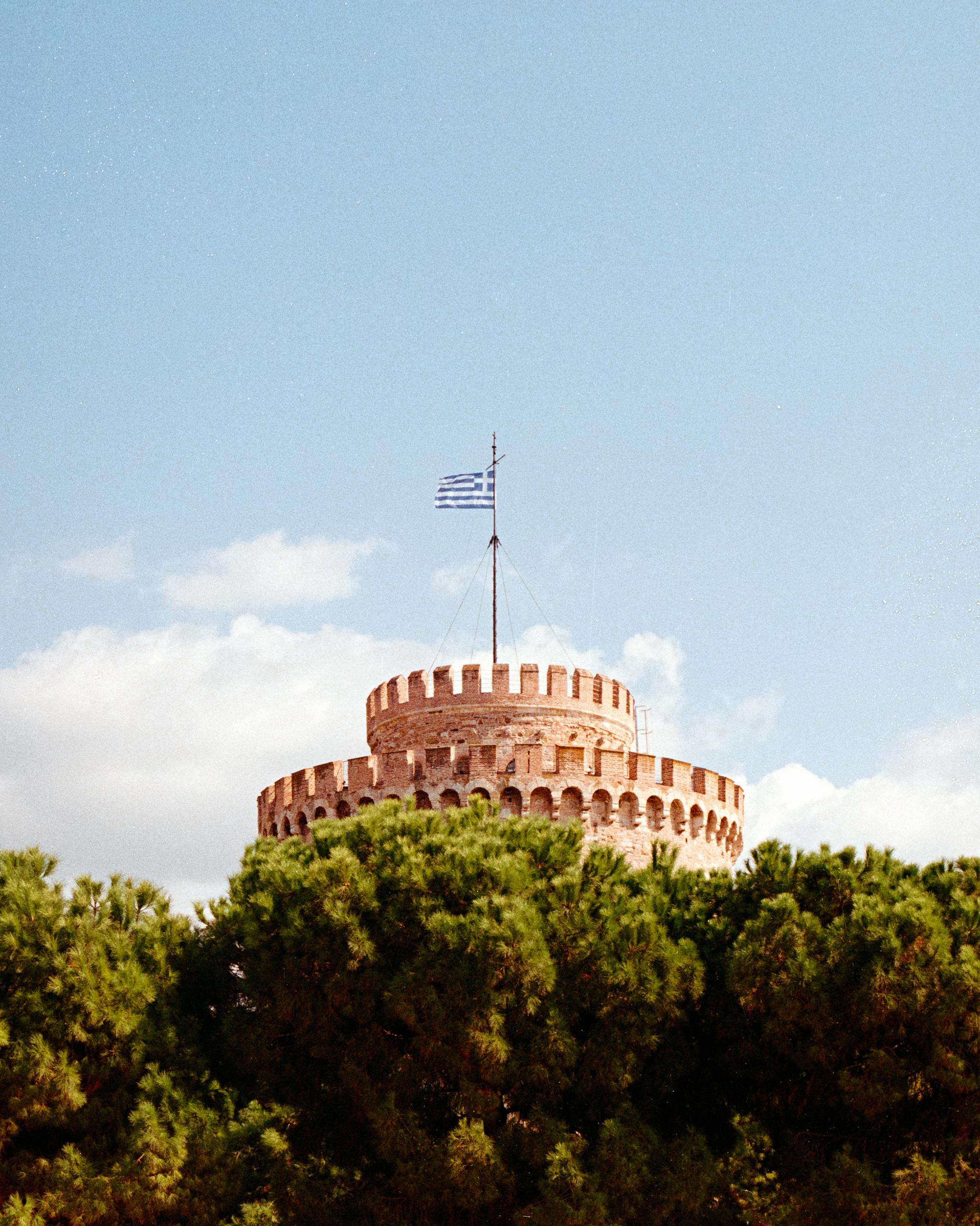 The historic White Tower of Thessaloniki with a Greek flag, under a bright blue sky.